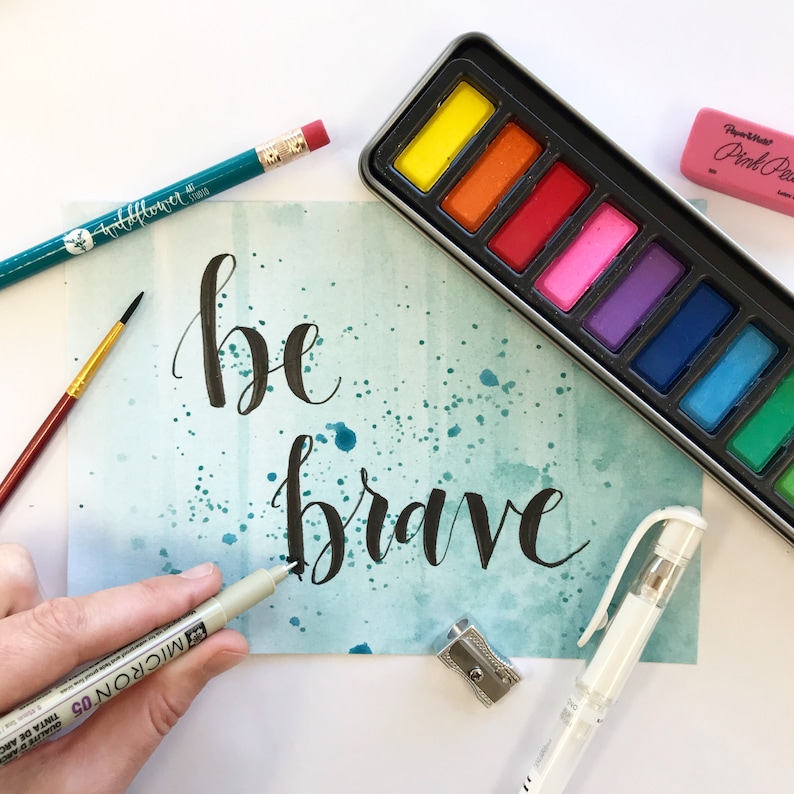 May include: A watercolor painting with a teal background and white splatters. The words "be brave" are written in black calligraphy. The image also includes a watercolor palette, a pencil, a pencil sharpener, and a pen.