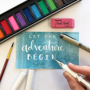 May include: A teal watercolor card with the text "Let the adventure begin" written in white. The card is surrounded by art supplies, including a set of watercolor paints, a pink eraser, and two white pens.