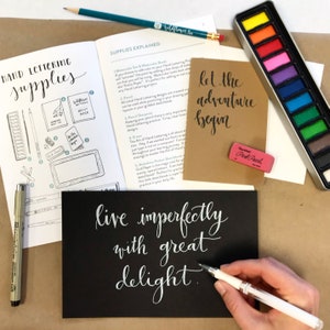 May include: A hand lettering kit with supplies including a watercolor set, a pencil, a pen, a small notebook, and a guidebook with the text "Hand Lettering Supplies" and "Supplies Explained". The guidebook includes instructions on how to use the supplies and create hand lettering designs. The kit also includes a black card with the text "live imperfectly with great delight" written in white calligraphy.