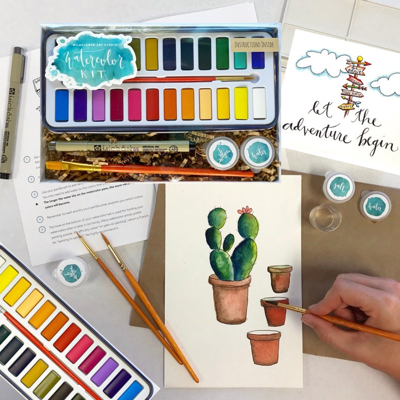 May include: A watercolor painting kit with 18 watercolor paints, two paintbrushes, a black pen, and a small container of water. The kit is open and the paints are visible. There is also a drawing of a cactus in a pot, with the words "Let the adventure begin" written on a piece of paper.