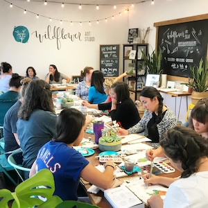 May include: A group of people are gathered around a table in a bright, airy studio, painting with watercolors. The studio is decorated with plants and a chalkboard sign that reads "Wildflower Art Studio".