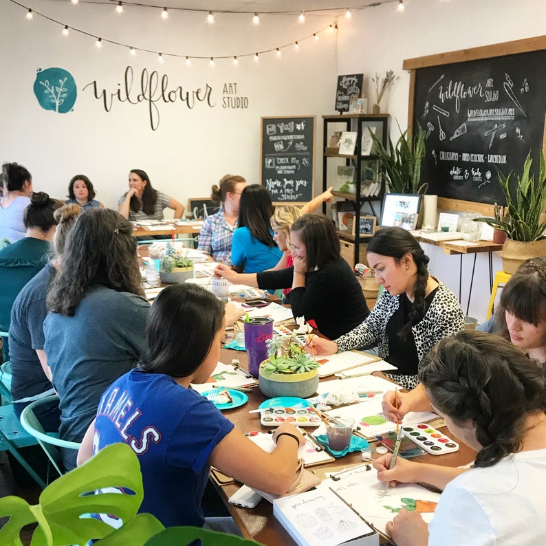 May include: A group of people are gathered around a table in a bright, airy studio, painting with watercolors. The studio is decorated with plants and a chalkboard sign that reads "Wildflower Art Studio".