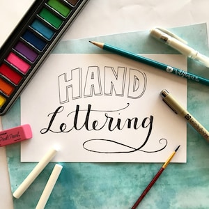 May include: A white sheet of paper with the words "HAND Lettering" written in black ink. The paper is surrounded by art supplies, including a watercolor palette, a pencil, a black marker, and a white eraser.