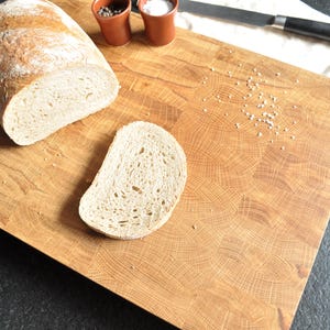 May include: A wooden cutting board with a loaf of bread and a slice. The board has a natural wood grain pattern. Small brown pots of salt and pepper are next to a knife on a white cloth.