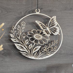 Könnte beinhalten: Wanddekoration aus Holz mit einem lasergeschnittenen Design eines Schmetterlings und Blumen in einem Kreis. Das helle Holz hebt sich von einem dunkelgrauen Hintergrund ab. Dieses dekorative Stück zeigt detaillierte Blumen- und Schmetterlingselemente, ideal für die Inneneinrichtung.