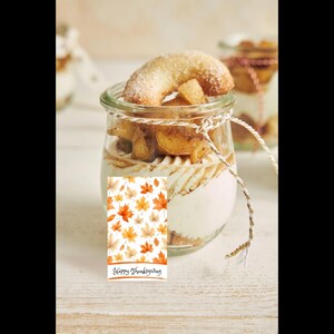 May include: A glass jar containing layers of cream, baked goods, and a crescent-shaped pastry dusted with icing sugar. A small card with orange and yellow autumn leaves and the text "Happy Thanksgiving" is tied to the jar.