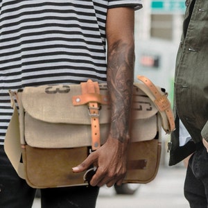 May include: A man wearing a striped black and white t-shirt with a red trim and a brown leather messenger bag with a canvas flap. He is standing in front of a building and talking to another man.