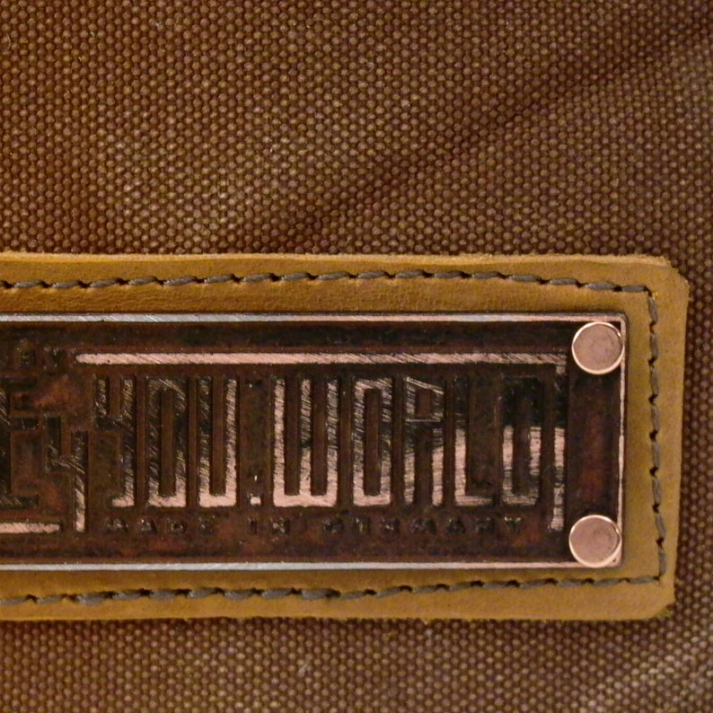 Puede incluir: Un parche de cuero marr&oacute;n con un borde cosido y una placa de metal de color cobre que dice "YOU.WORLD MADE IN GERMANY".