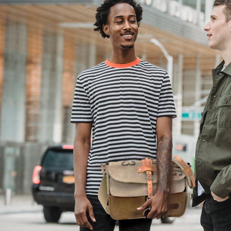 May include: A man wearing a striped black and white t-shirt with a red trim and a brown leather messenger bag with a canvas flap. He is standing in front of a building and talking to another man.