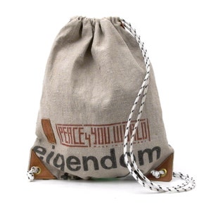 Könnte beinhalten: Eine beige Kordeltasche aus Leinen mit braunen Lederelementen. Die Tasche hat einen weißen und schwarzen Kordelzug und den Text "PEACE 4 YOU WORLD" in Braun auf der Vorderseite aufgedruckt. Der Text "MADE IN GERMANY" ist in kleinerer Schrift unter dem Haupttext aufgedruckt. Das Wort "eigendom" ist in großen Buchstaben auf der Unterseite der Tasche aufgedruckt.