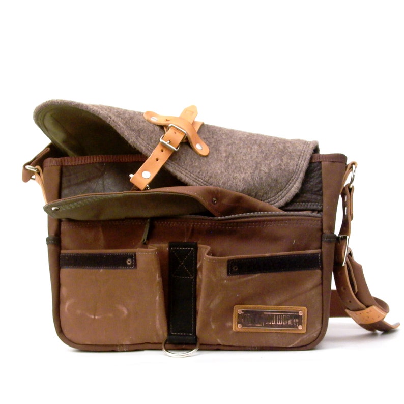 Puede incluir: Un bolso de mensajero de lona marr&oacute;n con m&uacute;ltiples bolsillos y una correa de cuero. El bolso tiene un parche de cuero marr&oacute;n con el texto "Field Notes" en &eacute;l. El bolso est&aacute; abierto y un forro de fieltro marr&oacute;n es visible en el interior.