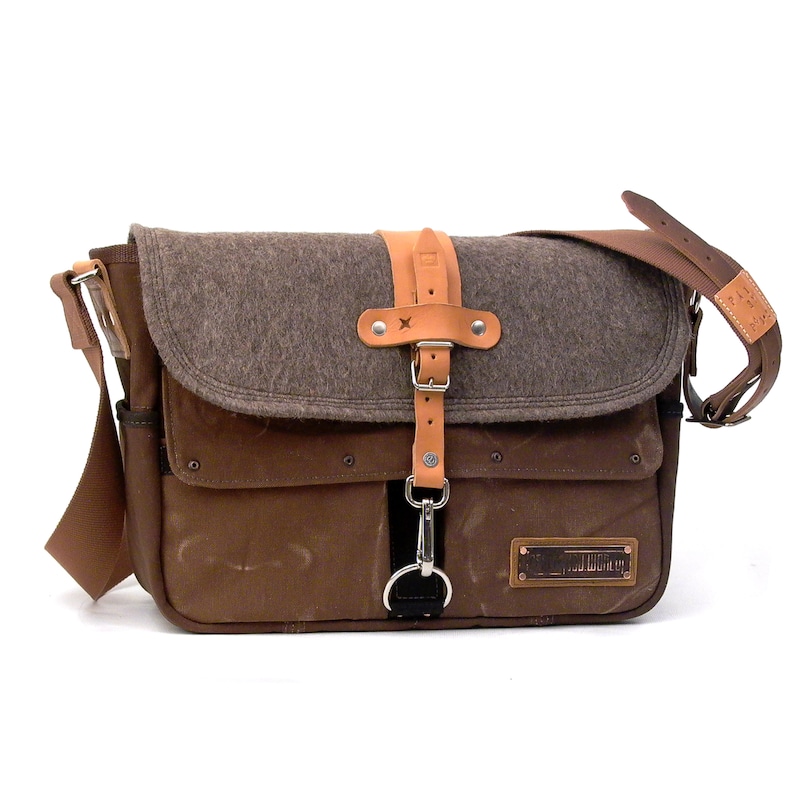 Puede incluir: Un bolso de mensajero de lona marr&oacute;n con una solapa de fieltro gris y detalles de cuero. El bolso tiene un cierre de metal y una correa de cuero con una hebilla de metal. El bolso tiene un parche de cuero marr&oacute;n con el texto "Field Notes" en &eacute;l.