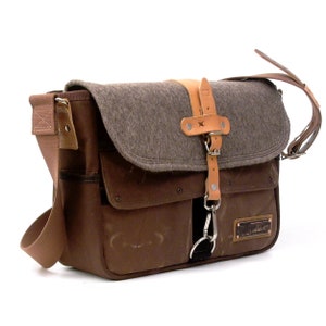 Puede incluir: Un bolso de mensajero de lona marr&oacute;n con una solapa de fieltro gris y detalles de cuero. El bolso tiene un bolsillo frontal grande y un bolsillo m&aacute;s peque&ntilde;o en el lateral. El bolso tiene una correa de cuero con hebilla y un clip de metal.