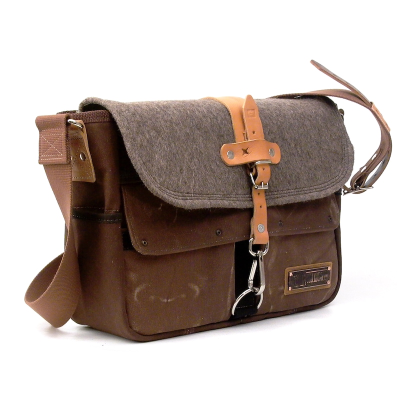 Puede incluir: Un bolso de mensajero de lona marr&oacute;n con una solapa de fieltro gris y detalles de cuero. El bolso tiene un bolsillo frontal grande y un bolsillo m&aacute;s peque&ntilde;o en el lateral. El bolso tiene una correa de cuero con hebilla y un clip de metal.