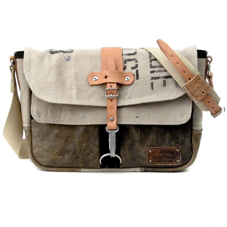 May include: A brown and beige canvas messenger bag with a leather strap and buckle. The bag has a brown leather patch with the text "Peace Love Unity" on it.