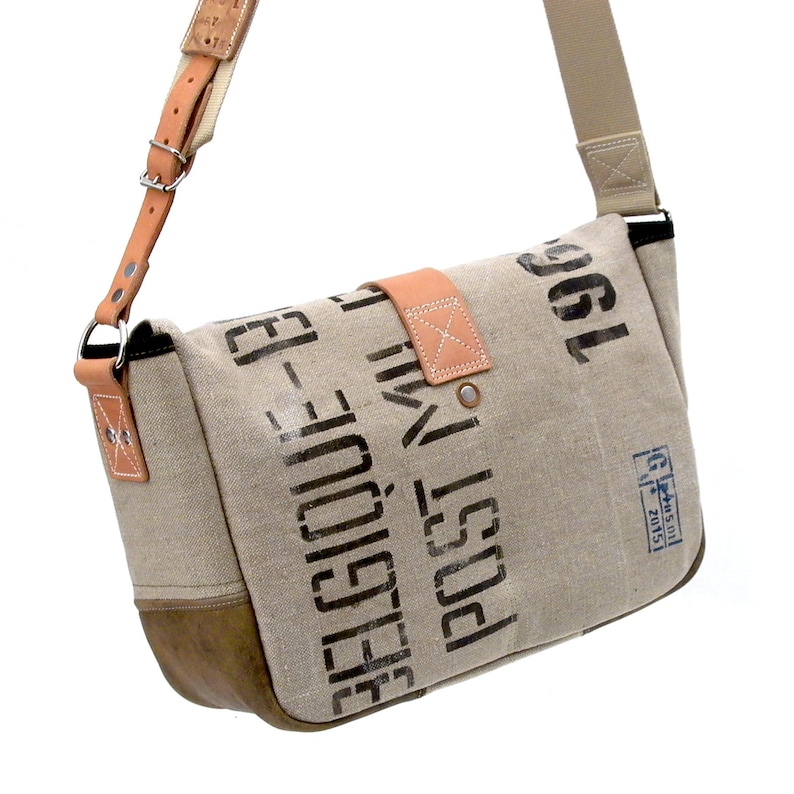May include: A beige canvas messenger bag with a brown leather flap and strap. The bag has black printed text that reads "Post" and "1961".