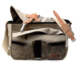 May include: A brown leather and canvas messenger bag with a large flap and multiple pockets. The bag has a brown leather strap and a metal buckle. The bag has a label that says "Remarkable Peace Movement".