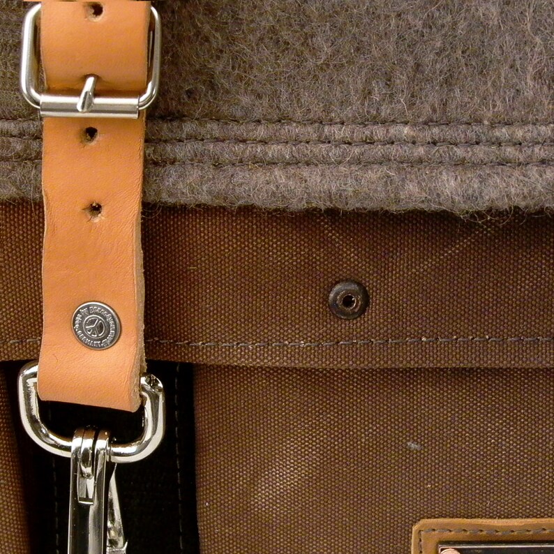 Puede incluir: Primer plano de una correa de cuero marr&oacute;n con una hebilla de metal y una bolsa de lona marr&oacute;n con un forro de fieltro gris. La bolsa tiene un remache de metal y detalles de costura.
