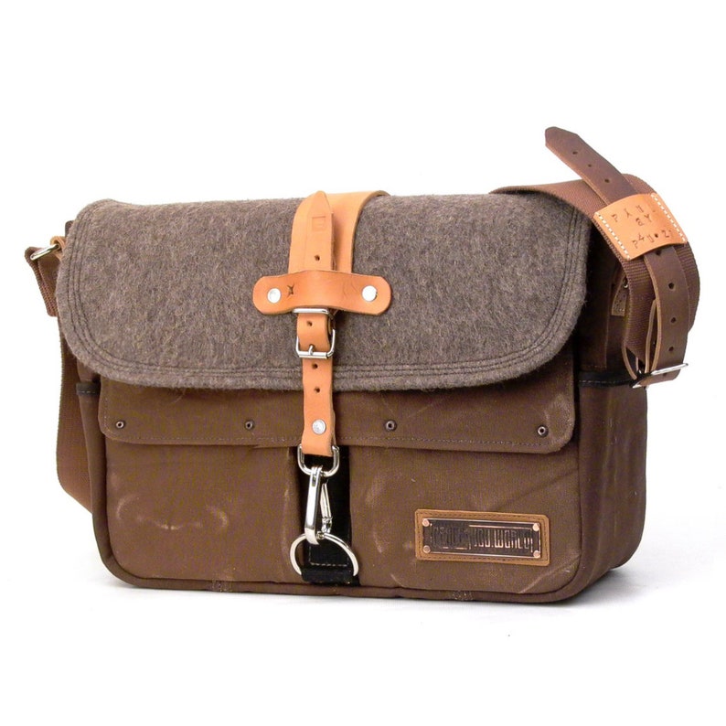 Puede incluir: Un bolso de mensajero de lona marr&oacute;n con una solapa de fieltro marr&oacute;n y una correa de cuero. El bolso tiene una hebilla de metal y un gancho de metal. El bolso tiene un parche de cuero marr&oacute;n con el texto "The  New  World" en &eacute;l.