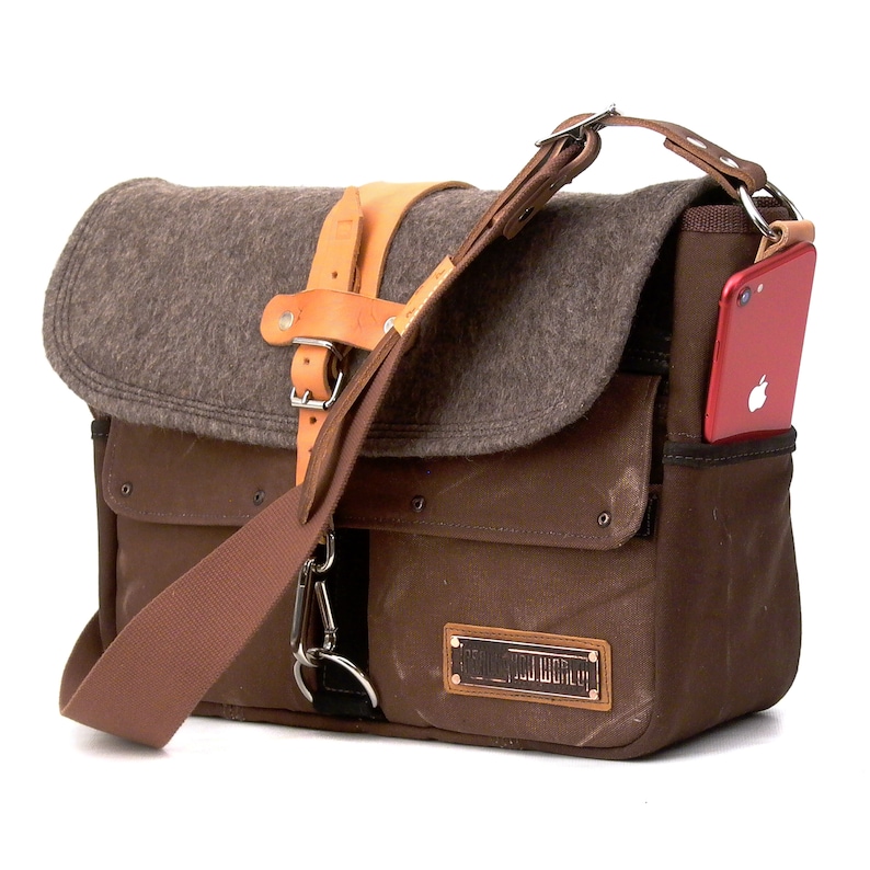 Puede incluir: Un bolso de mensajero de lona marr&oacute;n con una solapa de fieltro gris y una correa de cuero. El bolso tiene una hebilla de metal y un peque&ntilde;o bolsillo en la parte delantera. El bolso tiene una correa de cuero marr&oacute;n y una funda de tel&eacute;fono roja unida al lateral.