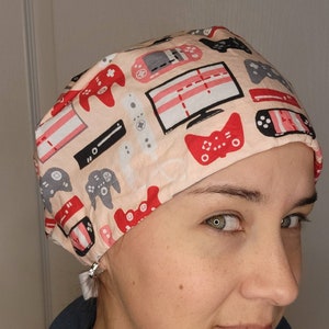 May include: A scrub cap featuring a light peach background with a repeating pattern of red, black, and grey video game controllers, handheld gaming consoles, and television screens displaying game graphics in a scattered, multi-directional layout.
