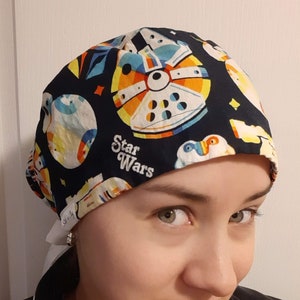 May include: A black scrub hat with a white tie. The hat has a colorful Star Wars print featuring the Death Star and the text "Star Wars".