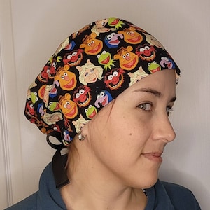 May include: A black scrub hat with a colorful print of various Muppet characters, including Kermit the Frog, Miss Piggy, and Animal. The hat has a tie that can be tied at the back of the neck.