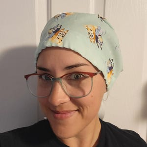 May include: A light blue surgical scrub cap with a repeating pattern of cartoon dogs in various poses. The cap is made of a soft fabric and is designed to cover the hair during medical procedures.