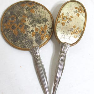 May include: Two antique vanity mirrors. One is round, the other oval. Both have gold-toned frames and silver-colored handles. The mirror surfaces feature a floral design in shades of gold and brown. The handles have an ornate, Art Deco style.