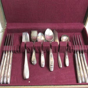May include: A set of silver-toned flatware including forks, spoons, and knives arranged in a rectangular storage case. The case features a deep burgundy velvet interior lining with molded slots to hold each piece of cutlery securely in place.