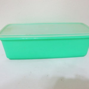 May include: A rectangular, turquoise plastic food storage container with a clear, rectangular lid. The container is empty and sits on a white surface. The lid is slightly translucent, allowing a glimpse of the container's contents.