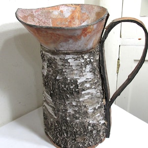 May include: A handcrafted pitcher with a unique design. The body is made from birch bark, with a textured, natural appearance. The top has a rusty orange and brown color, and the handle is made from a dark, curved branch. A decorative piece.