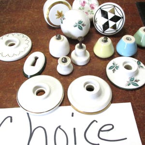 May include: A collection of vintage ceramic door knobs in various colors and patterns, including white, green, yellow, blue, and black. Some knobs feature floral designs, while others have geometric patterns. The knobs are arranged on a wooden surface with a handwritten sign that says "Choice".