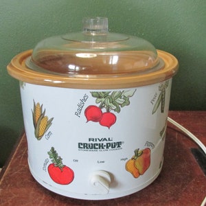 May include: A white Rival Crock-Pot slow cooker with a clear glass lid and tan trim. The slow cooker has illustrations of vegetables and the text "Rival Crock-Pot". It has a white power cord and a three-prong plug.