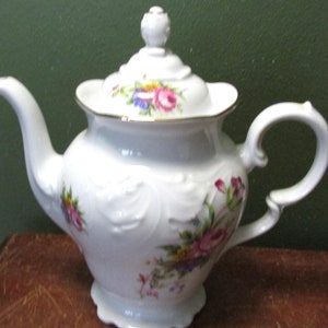 May include: White porcelain teapot with a floral design. The teapot has a lid with a knob on top and a handle on the side. The teapot is decorated with pink, purple, and yellow flowers.