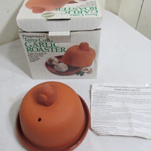 May include: A terra cotta garlic roaster with a lid and base, displayed with its original packaging. The box reads "Progressive Terra Cotta Garlic Roaster". The roaster is designed for roasting garlic or onions.
