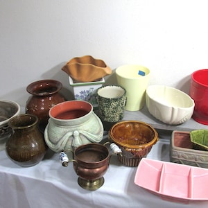 May include: A collection of 13 ceramic and metal planters in various shapes, sizes, and colors. The planters include a green planter with a bow, a red planter, a brown planter with a textured surface, a white planter with a floral design, and a copper planter with a brass base.
