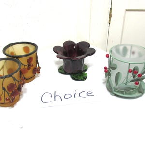 May include: A collection of decorative candle holders. Two amber-colored glass holders with a berry design, a dark purple flower-shaped holder, and a green metal holder with red berries and leaves. The word "Choice" is written on a white paper.