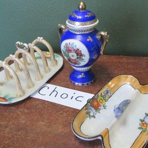 May include: A collection of vintage porcelain serving pieces. Includes a blue and gold lidded urn with floral details, a divided dish with a blue bird, and a toast rack. The word "Choice" is visible.