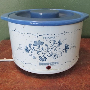 May include: A white Rival Crock-ette slow cooker with a blue lid and trim. The cooker features a floral design in blue and the text "RIVAL" and "Crock-ette". A red light indicates the cooker is on.
