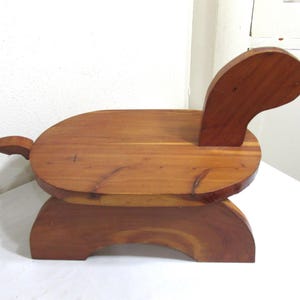 May include: A small wooden stool crafted from reddish-brown wood with a smooth, polished finish. The stool features a flat, oval-shaped seat, a curved upright backrest, a small tail-like protrusion at the rear, and a sturdy arched base.