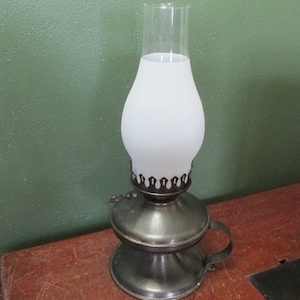 May include: A vintage oil lamp with a frosted glass globe and a clear glass chimney. The lamp has a metal base and a handle. The lamp is sitting on a wooden surface. The background is a solid green color.