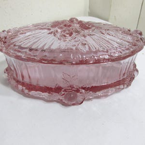 May include: An oval-shaped pink glass trinket box featuring a removable lid with a raised floral and leaf pattern. The sides of the container are smooth with a scalloped base edge, while the lid displays a textured design of roses and foliage.