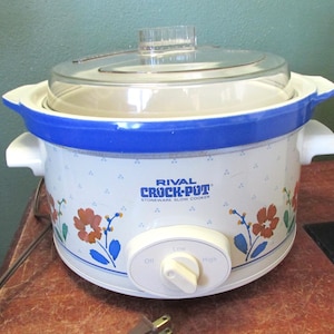 May include: A white Rival Crock-Pot stoneware slow cooker with a clear glass lid. The slow cooker has a blue rim and handles, with floral and leaf designs. The control knob has settings for Off, Low, and High.