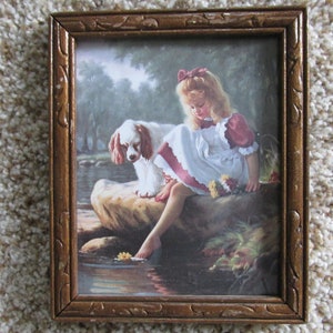 May include: A framed print depicts a young person in a white dress with red accents, sitting by a stream with a Cavalier King Charles Spaniel. The print has a gold-colored, ornate frame. The scene is set in a natural outdoor environment.