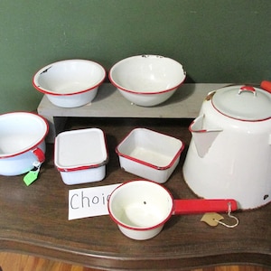 May include: A collection of vintage white enamelware kitchen items with red trim. Includes bowls, a kettle, and containers. The items are arranged on a wooden surface, showcasing a rustic aesthetic. Some pieces show signs of wear.