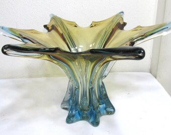 May include: A decorative glass bowl with a starburst design. The bowl features a combination of colors, including amber, blue, and brown. The bowl has a wide, open top and a pedestal base. The glass has a glossy finish.