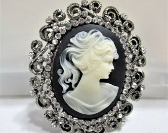 May include: A vintage-style cameo brooch featuring a profile of a woman in ivory against a black oval background. The cameo is set within a silver-toned filigree frame, embellished with clear rhinestones. A classic jewelry piece.
