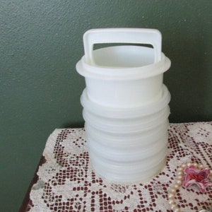 May include: A white plastic container with a handle, stacked with several circular sections. The container is sitting on a white lace doily. A pearl necklace with a pink flower is in the foreground.