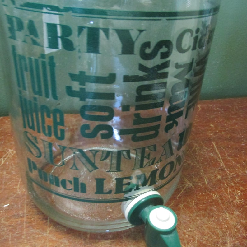 May include: A clear glass beverage dispenser featuring dark green text printed on the exterior. The text includes words like PARTY, fruit juice, soft drinks, SUN TEA, and LEMON. A green and white plastic spigot is attached to the lower front side.